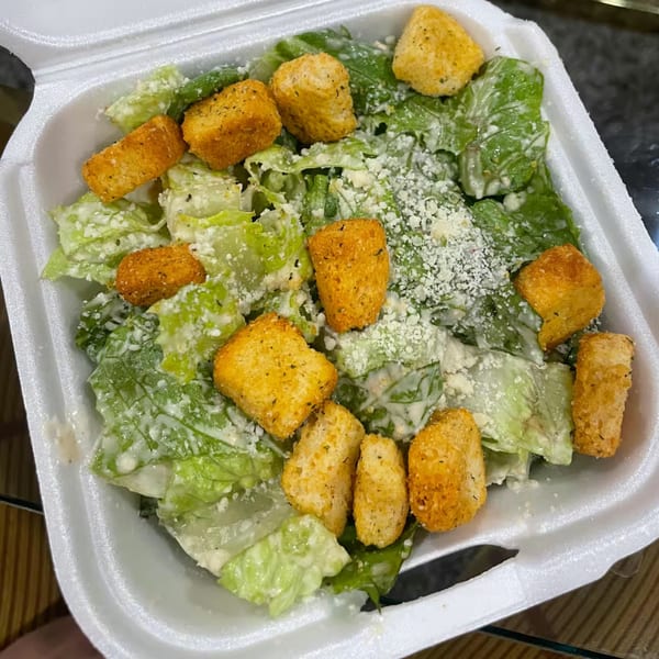 Caesar salad with romaine lettuce, creamy dressing, grated cheese, and golden croutons in a white takeout container.